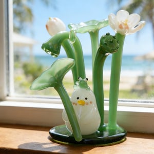 May include: A whimsical glass sculpture featuring a white chick with a yellow beak and rosy cheeks, nestled among green lily pads and stems. The sculpture includes a frog and a white flower with pink accents. The base is dark green.
