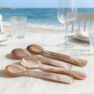 Puede incluir: Cuatro cucharas de servir nacaradas en tonos marrón claro y crema se colocan sobre un mantel blanco. Las cucharas tienen un acabado liso y brillante y están cerca de un vaso de agua y una copa de vino. El fondo muestra el mar.