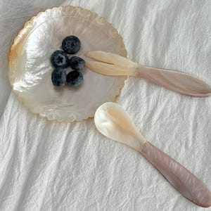 May include: A pearlescent serving set with a scalloped edge plate, a spoon, and a fork. The spoon and fork have pink handles. The plate holds several blueberries. The set is on a white, textured surface.