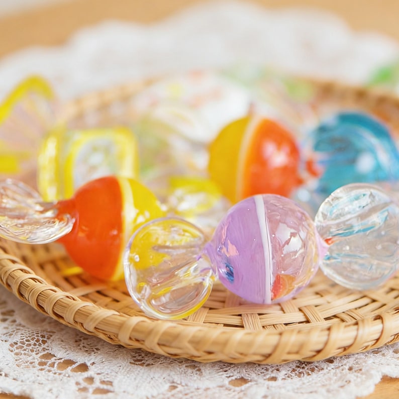 May include: A collection of colorful, glass-blown candies in a woven basket. The candies feature vibrant hues of orange, yellow, purple, and blue, with clear, twisted glass accents. The basket sits on a white lace doily.