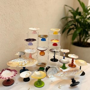 May include: A collection of decorative glass and shell serving dishes in various colors and designs. Some dishes are displayed on clear acrylic stands. Pearls are scattered on the white surface.
