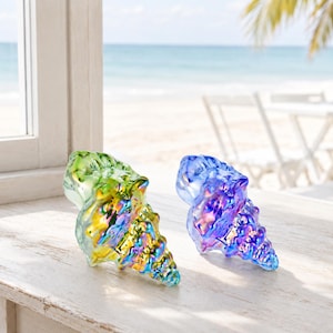 May include: Two decorative seashell figurines made of iridescent glass. One shell features a vibrant blend of green and yellow hues, while the other displays a deep blue and purple gradient. Both items have a glossy, reflective finish.