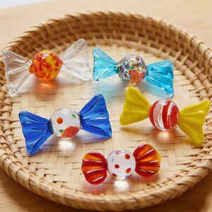 May include: Five colorful glass candy ornaments. Each ornament features a round, speckled center with clear, blue, yellow, or orange bow-shaped accents. The ornaments are displayed in a woven basket.
