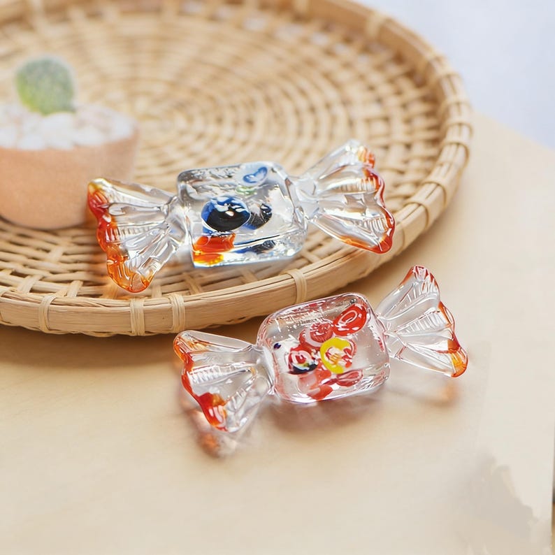 May include: Two clear glass candy-shaped ornaments with colorful details. The ornaments have orange and red accents on the edges. One ornament has blue, black, and orange details, while the other has red, yellow, and orange details.