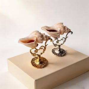 May include: Two conch shell decorative pieces. One shell is mounted on a gold-colored metal stand with floral accents, and the other is on a silver-colored metal stand. The shells are a light peach color, and the stands are on a beige surface.