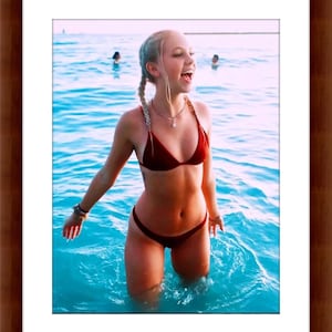 May include: A framed photograph shows a person in a burgundy bikini standing in turquoise water. The person is smiling with their mouth open. Other people are visible in the background, in the water. The photograph is framed in a dark brown wooden frame.