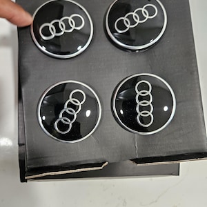 May include: Four black and silver Audi wheel center caps are displayed in a cardboard box. Each cap features the Audi logo, a set of four interlocking rings, in silver against a black background.