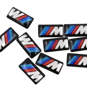 Pode incluir: Uma coleção de emblemas retangulares pretos com as letras brancas "M" e "W" e listras vermelhas, azuis e brancas. Os emblemas estão dispostos sobre um fundo branco.