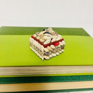 May include: A square trinket box covered in small, assorted seashells in shades of white, tan, and brown. The lid features a dense cluster of shells, while the sides are decorated with rows of shells. A thin red trim is visible around the lid edge.