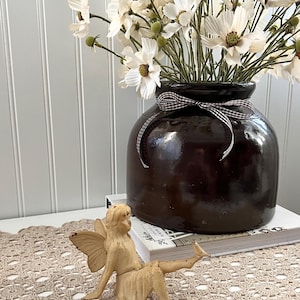 May include: A brown ceramic vase with a black and white checkered ribbon holds white flowers. A small, tan-colored angel figurine sits on a beige crocheted table runner. A book is visible beneath the vase.