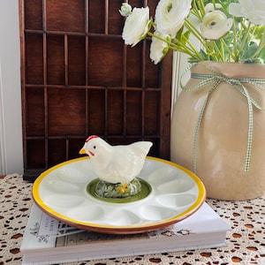 May include: A white ceramic egg holder with a yellow rim and a decorative white chicken figurine. The egg holder sits on a stack of books, with a vase of white flowers in the background.