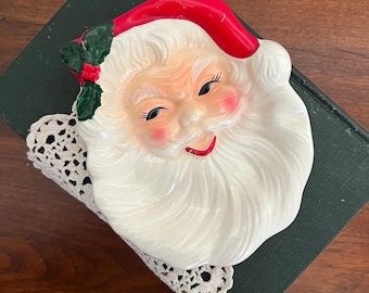 May include: A ceramic Santa Claus face-shaped dish with a red hat, white beard, and rosy cheeks. The dish is decorated with holly and rests on a white lace doily and a dark green book. A festive Christmas decoration.
