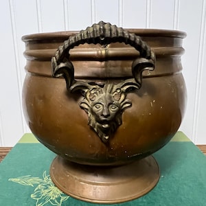 May include: A round copper pot with a wide rim and a pedestal base. The pot features a prominent dark metal handle attached to a decorative relief of a stylized face with horns, centered on the front of the vessel's exterior surface.