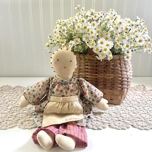 May include: A fabric doll with a cream-colored head, simple stitched facial features, and red hair accents. The doll wears a floral print long-sleeve dress, a plain cream apron embroidered with the name Esther, and red plaid trousers.