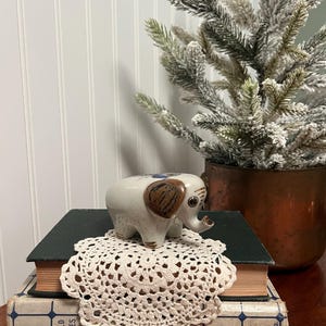 May include: A ceramic elephant figurine with brown ears and blue accents sits atop a stack of vintage books and a cream-colored doily. A small, decorative Christmas tree is in the background.