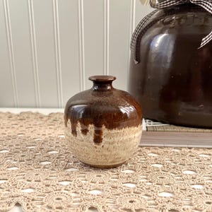 May include: A small, round, brown ceramic vase with a glossy finish. The top is a dark brown, transitioning to a lighter, speckled beige at the base. The vase is displayed on a beige crocheted doily, with a larger brown jug in the background.