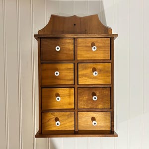 May include: A wall-mounted wooden storage cabinet featuring eight rectangular drawers arranged in two vertical columns. Each drawer is crafted from medium-toned wood and is equipped with a small, round, white ceramic knob centered on the front.
