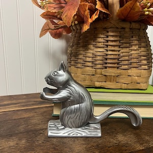 May include: A silver-toned metal nutcracker shaped like a squirrel sitting on a log base. The squirrel has a textured body, pointed ears, and a long curved tail that functions as a lever. The metal surface has a smooth, matte finish.