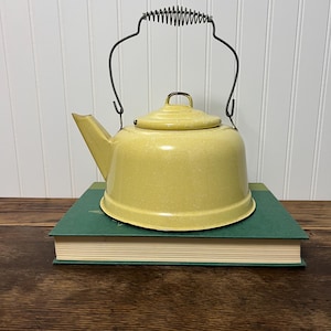 May include: A pale yellow enamel tea kettle with a speckled finish, featuring a matching lid and a dark metal wire handle with a coiled grip. The kettle has a wide base and a short, angled spout, resting on a thick, dark green hardcover book.