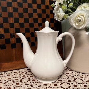May include: A white ceramic teapot with a lid, spout, and handle. The teapot has a bulbous body and a decorative floral design on the lid. The teapot is on a cream-colored lace tablecloth. A vase of white flowers is in the background.