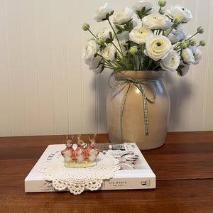 May include: A decorative arrangement featuring a beige ceramic vase with white flowers and a green ribbon. A small figurine of three rabbits in red coats sits on a book atop a white lace doily. The book is titled "Liz Marie Galvan".