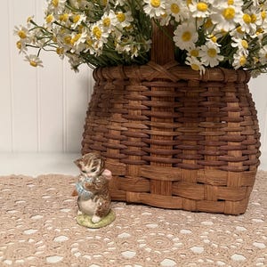 May include: A small ceramic figurine of a brown tabby kitten holding a blue ball of yarn, positioned in front of a woven brown wooden basket filled with a large bouquet of white and yellow daisy-like flowers on a beige crocheted lace mat.