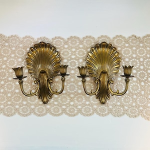 May include: A pair of matching brass wall sconces, each featuring a central shell-shaped backplate with vertical ridges and two curved arms extending outward. Each arm terminates in a small, tulip-shaped candle cup holder for taper candles.