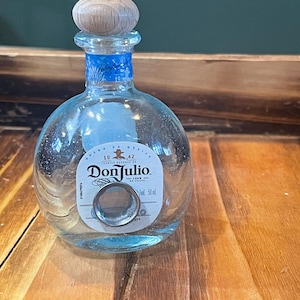 Może przedstawiać: Mała, przezroczysta butelka tequili Don Julio z drewnianym korkiem. Butelka ma niebieską etykietę z białym tekstem i srebrnym pierścieniem. Butelka ma 50 ml.