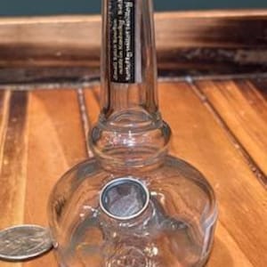 May include: A clear glass bottle with a round base and a long, slender neck. The bottle has a small, circular opening near the base and a black label around the neck. A US quarter is next to the bottle. The bottle is sitting on a wooden surface.