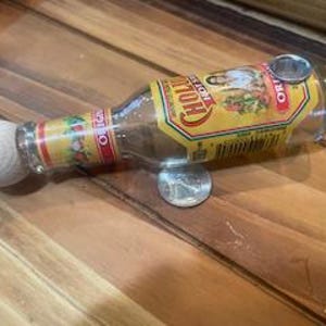 Op de afbeelding: Een hergebruikte glazen fles met een geel en rood etiket, voorzien van een witte, bolvormige stopper en een metalen pijpaansluiting. De fles ligt op een houten oppervlak, ondersteund door een munt.