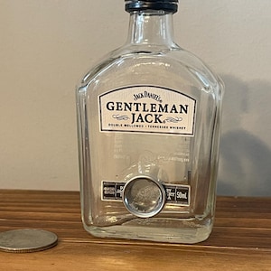 May include: A clear, empty 50ml Gentleman Jack whiskey bottle with a black cap. The label reads "Jack Daniel's Gentleman Jack Double Mellowed Tennessee Whiskey". A silver coin sits on the wooden surface.