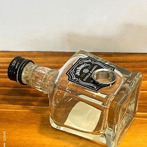 May include: A clear glass bottle with a black cap and a label that reads "Jack Daniel's Single Barrel." The bottle has a square base and a rounded neck. A metal screen is visible inside the bottle.