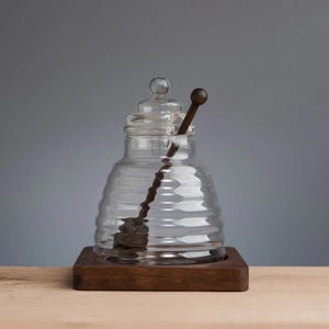 Könnte beinhalten: Ein klarer Honigtopf aus Glas mit Deckel und einem Honiglöffel aus Holz. Der Topf hat eine Bienenwabenform und steht auf einem kleinen, quadratischen Holzsockel. Der Löffel ist dunkelbraun und liegt im Glas.