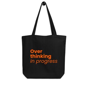 Könnte beinhalten: Schwarze Tragetasche mit dem orangefarbenen Text "Over thinking in progress". Die Tasche hat lange Henkel und ist aus robustem Material gefertigt. Die Tasche hängt an einem schwarzen Haken.