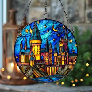 Könnte beinhalten: Rundes Buntglasfenster mit einer Burgszene unter einem Nachthimmel. Die Burg mit ihren Türmen wird von gelben und orangen Lichtern beleuchtet. Der blaue Himmel zeigt einen Mond und Sterne. Das Fenster ist von dunklem Metall umrandet und an einer Kette aufgehängt.