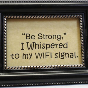 May include: A framed print with the text "Be Strong, I Whispered to my WIFI signal." The frame is black with a silver inner border and a textured design. The background of the print is a light tan color.
