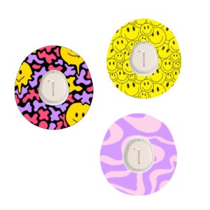 May include: Three Dexcom G6 sensor covers with colorful designs. One features a yellow and pink pattern with smiley faces, another has a yellow smiley face pattern, and the third has a pink and purple swirl design. Each cover has a white sensor.