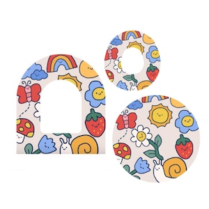 Puede incluir: Tres pegatinas troqueladas con un diseño caprichoso. Las pegatinas presentan un arcoíris, un sol, una mariposa, una flor, una fresa, una seta y un caracol sobre un fondo beige. Una pegatina es arqueada, otra circular y otra es un círculo con un agujero en el medio.