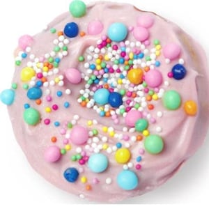 Puede incluir: Primer plano de una magdalena con glaseado rosa, cubierta con chispas y bolitas de caramelo de colores. Las chispas son blancas, amarillas, naranjas, verdes y azules. Las bolitas son rosas, azules, verdes y amarillas.