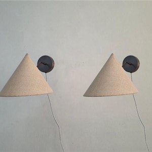 Puede incluir: Dos apliques de pared con pantallas cónicas de tela beige. Cada aplique tiene una base circular oscura y un cable. Las lámparas están montadas en una pared clara, proporcionando una luz suave y ambiental. Las pantallas miden unos 30 cm de diámetro.