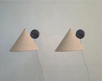 May include: Two wall-mounted sconces with conical, beige fabric shades. Each sconce has a dark, circular base and a wire extending from the shade. The lamps are mounted on a light-colored wall, providing a soft, ambient light.