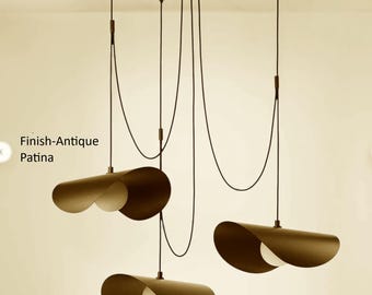 May include: A multi-light pendant chandelier with an antique patina finish. The fixture features three curved, dark-toned metal shades suspended by black cords from a central ceiling mount, each housing a single round white light bulb.
