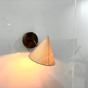 Puede incluir: Un aplique de pared con una base circular de tono bronce y una pantalla cónica hecha de tela texturizada de color beige claro. La pantalla está inclinada hacia abajo y emite un brillo cálido y suave que ilumina el área circundante.
