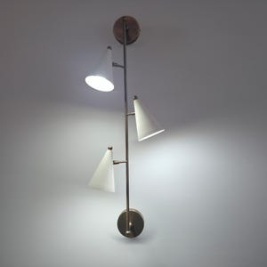 May include: A wall-mounted light fixture featuring a vertical bronze-toned metal rod with three conical white shades. The fixture includes two circular bronze wall plates at the top and bottom, with the three lights emitting a bright glow.