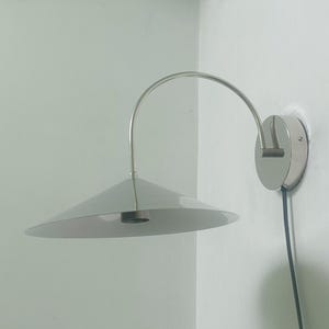 Peut inclure: Une applique murale moderne avec une finition argentée. La lampe présente un bras incurvé et un abat-jour conique. La lampe est fixée sur une base ronde argentée. Un cordon noir s'étend de la base.