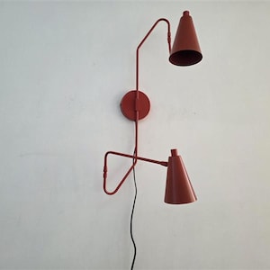Puede incluir: Una lámpara de pared roja con dos pantallas cónicas. La lámpara tiene una base circular y brazos angulados. Las pantallas apuntan hacia abajo. Un cable de alimentación negro cuelga debajo.