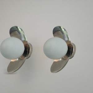 May include: A pair of wall sconces featuring a spherical white frosted glass shade centered in front of a curved, polished silver-tone metal backing. Each light fixture has a circular base plate and a reflective, wing-like metallic structure.