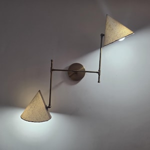 Puede incluir: Aplique de pared moderno con dos pantallas triangulares. El aplique tiene un acabado de latón y una base de pared circular. Las pantallas beige emiten una luz cálida, creando una luminaria elegante y funcional.