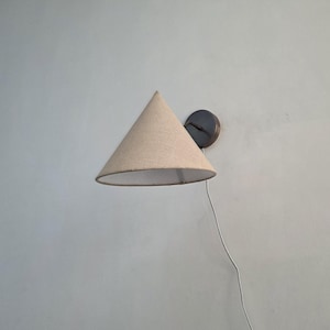 Può includere: Un'applique da parete con un paralume conico beige. Il paralume è fissato a una base circolare scura. Un cavo di alimentazione bianco si estende dall'apparecchio. La luce è progettata per uso interno.