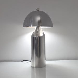May include: A polished silver-tone metal table lamp featuring a cylindrical base that tapers toward a conical neck, topped with a smooth, rounded dome-shaped shade. The lamp emits a soft, bright light from beneath the dome, and includes a black cord.
